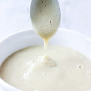 Creamy vegan cauliflower alfredo sauce dripping from a spoon into the bowl with more of it below.