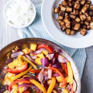 Mango tofu stir fry in a skillet next to a bowls of rice and jerk tofu.