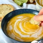 Hand dunking bread into creamy butternut squash soup with a swirl of coconut milk on top, another bowl of soup is in the background along with lime wedges and ginger.