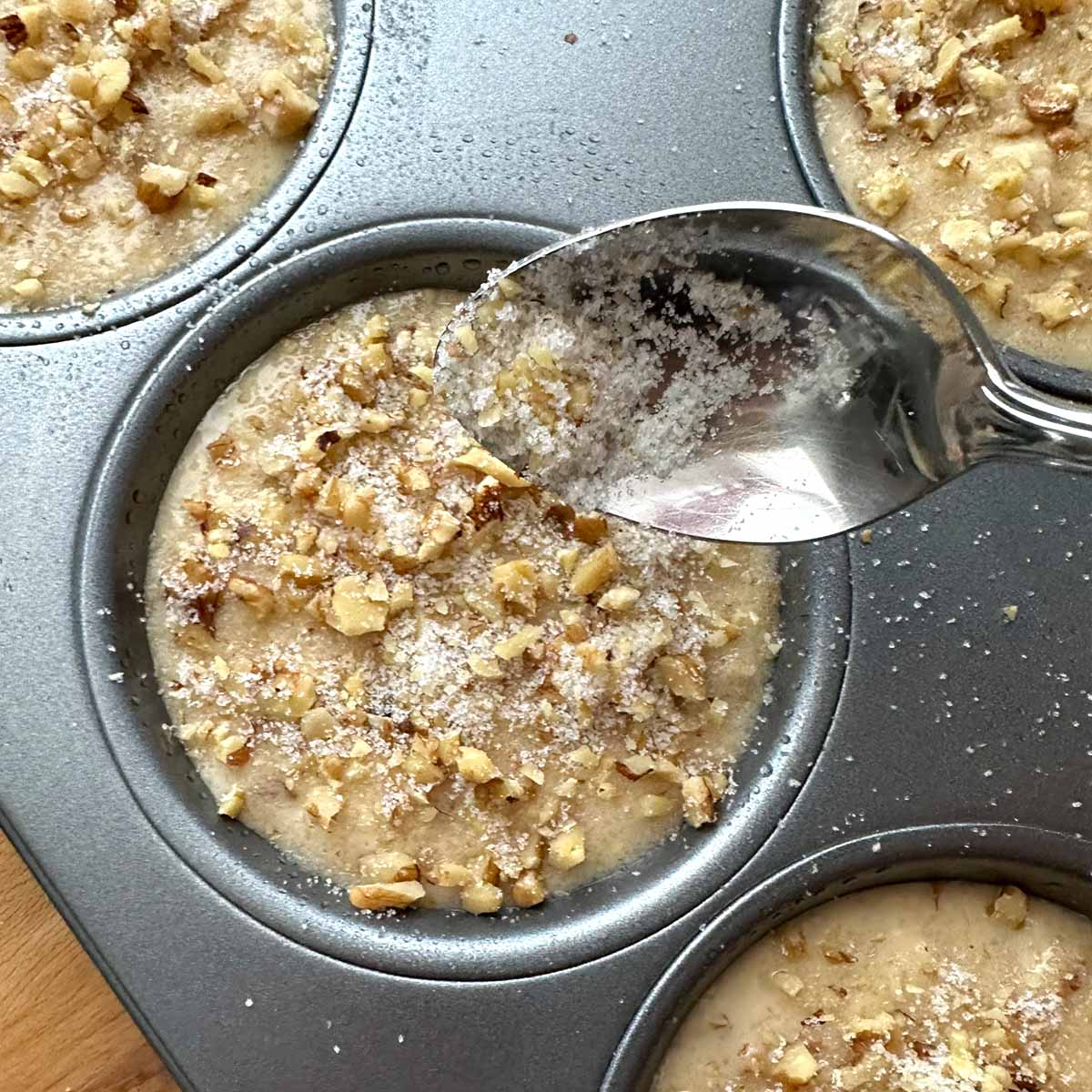 Muffin batter in tin with spoon sprinkling the nut and sugar topping on top.