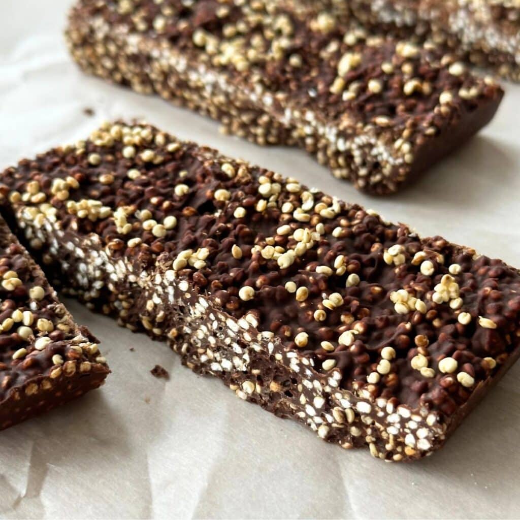 Chocolate quinoa crunch bar on sheet of parchment paper. In background are 2 more bars. Quinoa crunch bar is sliced through and the popped quinoa looks like tiny polka dots against dark chocolate.