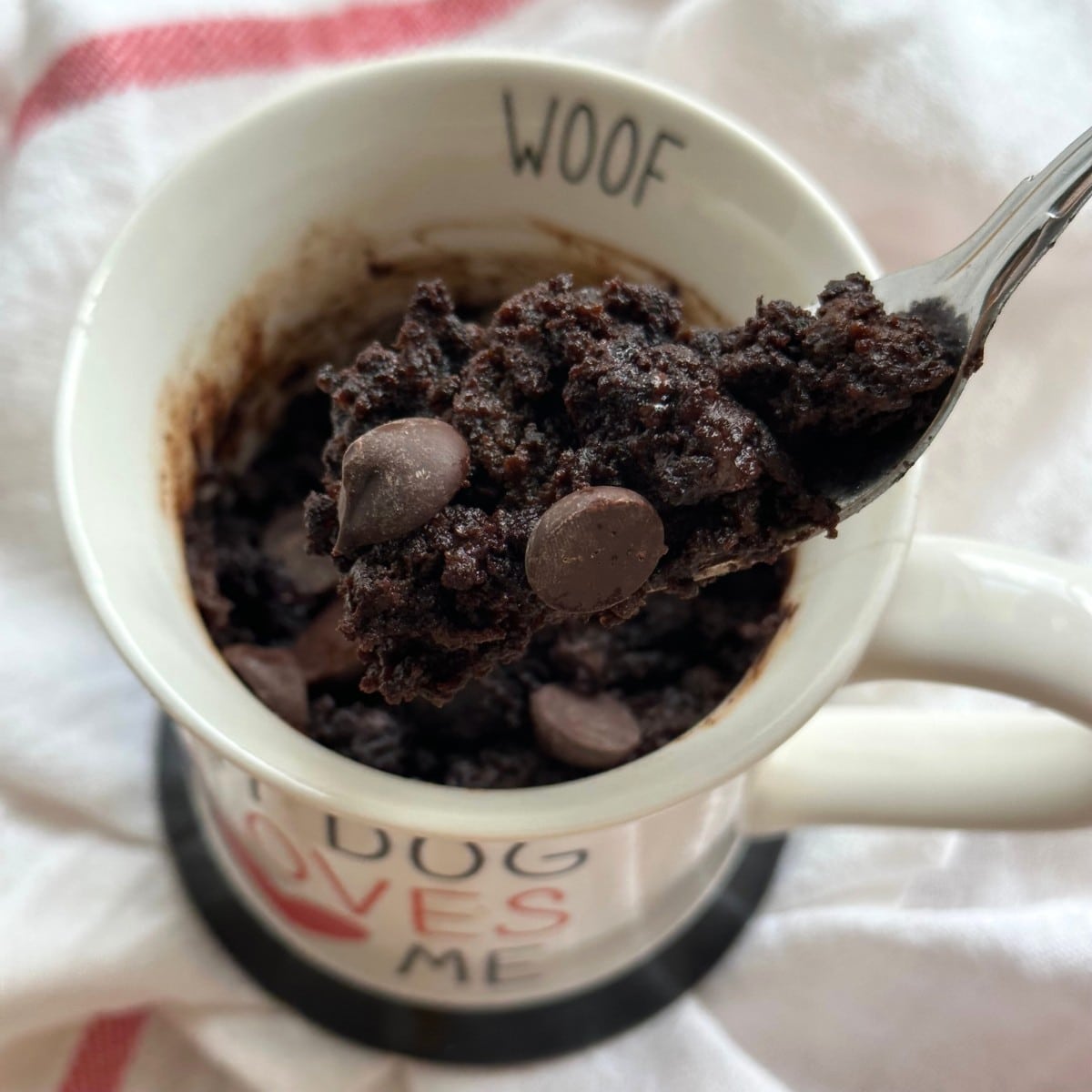 Bite of chocolate zucchini mug cake on fork lifted out of mug with more cake. White mug with text that reads "WOOF" on lip. On top of white cloth napkin with red stripes.