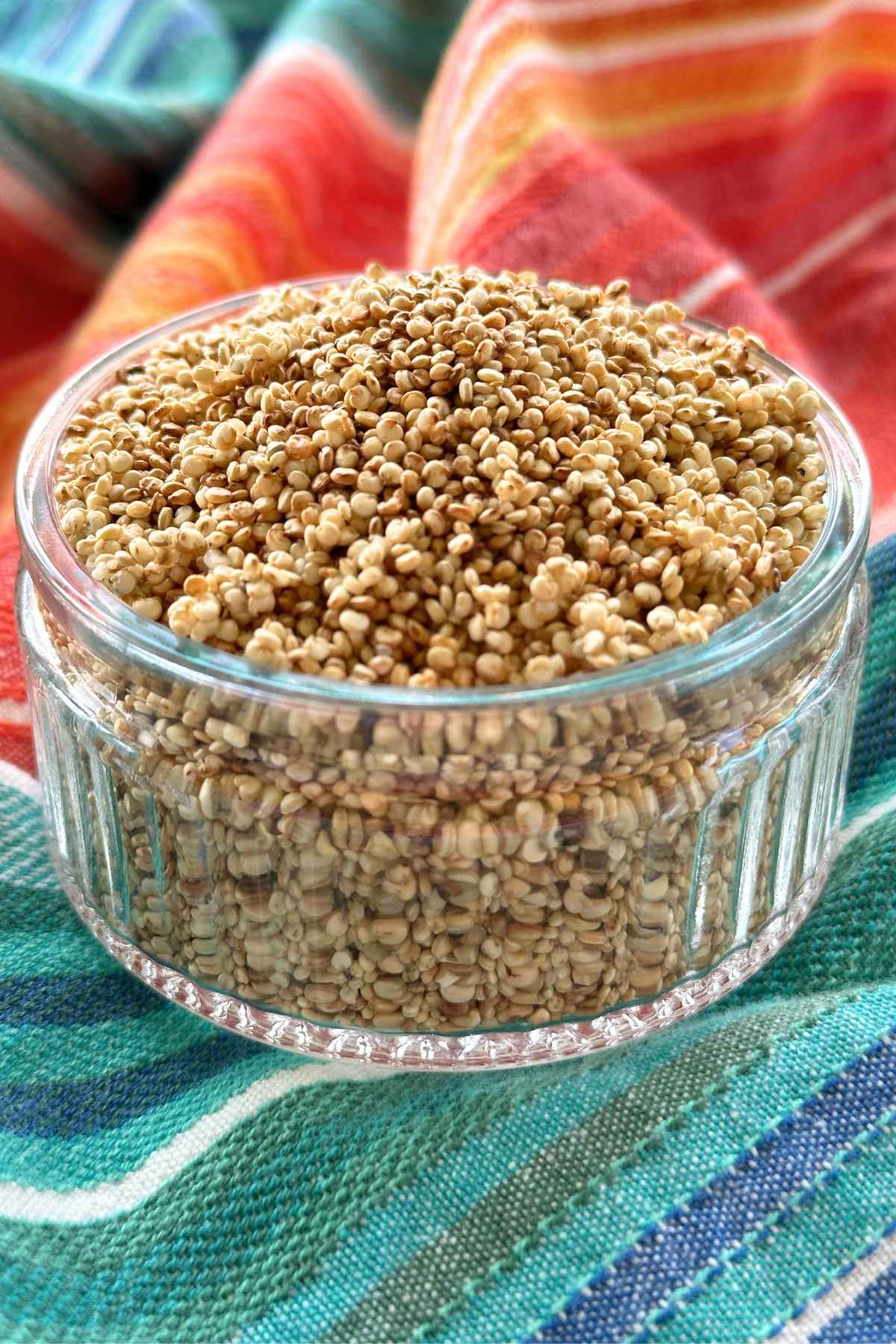 Glass bowl filled with golden colored popped quinoa seeds on red, orange, and blue cloth napkin in background.
