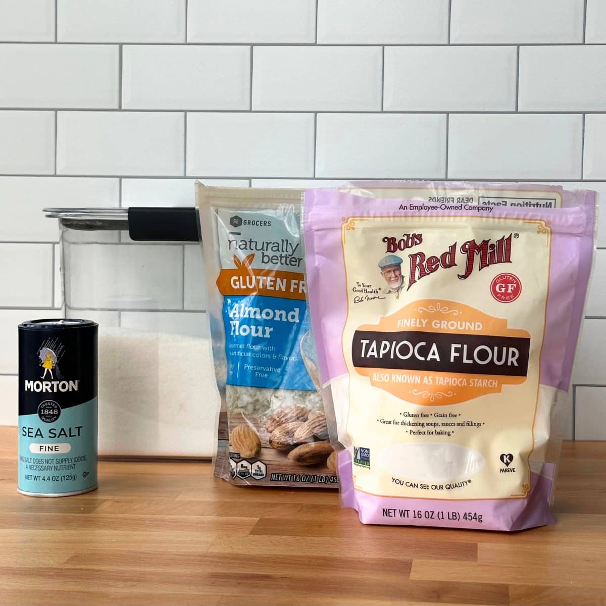 Ingredients to make gluten free vegan pie crust: fine sea salt, sugar, almond flour, and tapioca flour. Ingredients are on a wood surface with white subway tile wall in back.