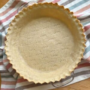Gluten free vegan pie crust parbaked in glass pie dish. Edges of the pie crust are crimped and golden in color. Pie is docked, has holes made with a fork, to allow steam to escape. The almond flour pie crust is on top of a white, pink, brown, and blue striped cloth napkin.
