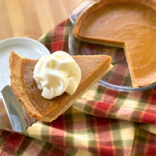 Slice of vegan sweet potato pie with dollop of whipped cream on top. Background shows the rest of the pie in a dish, a white plate, and a fall colored gingham napkin.