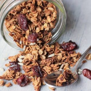 Maple vanilla granola spilling out of glass jar onto wood background with spoon. Granola is golden brown with cranberries and sunflower seeds.