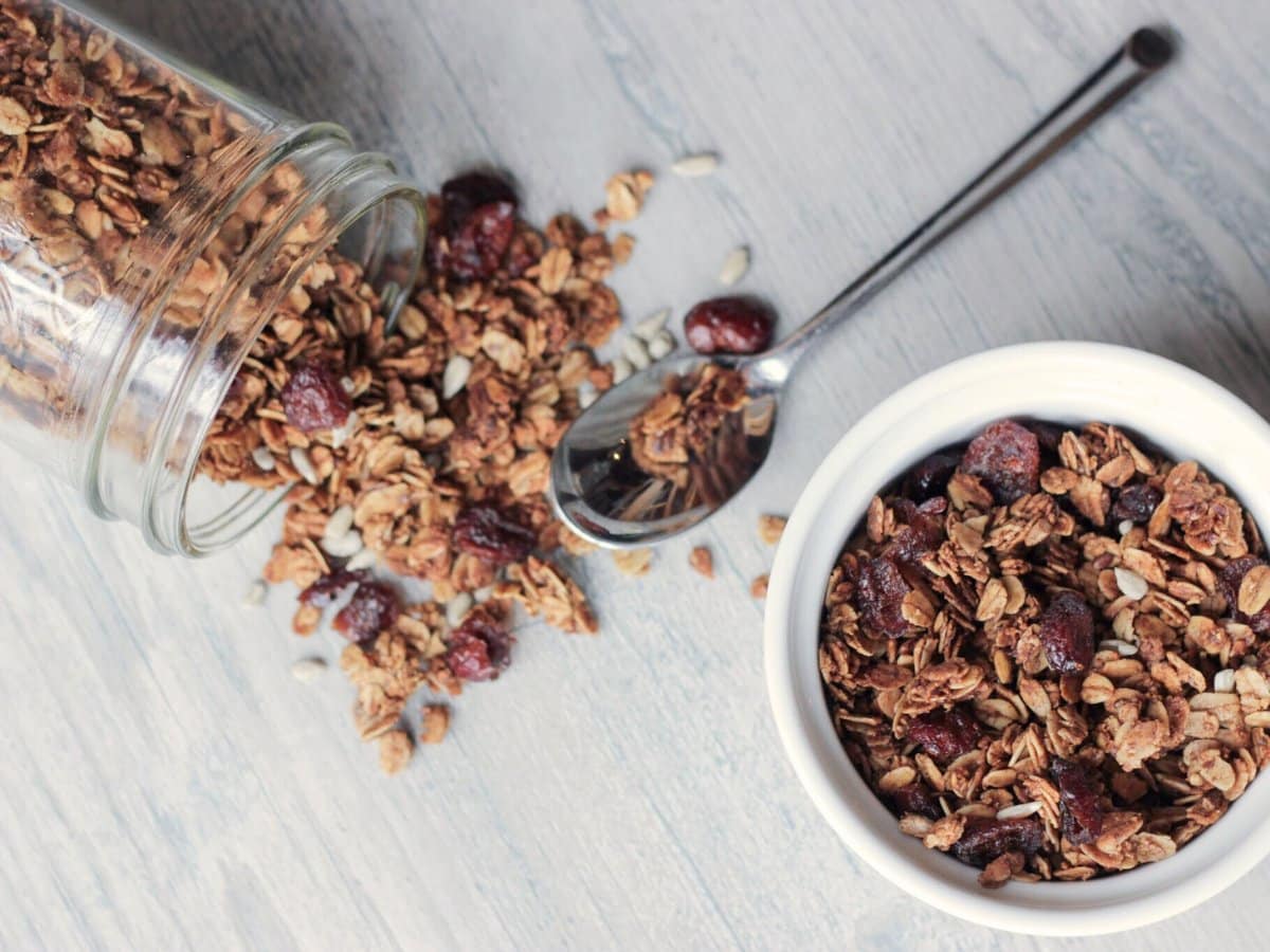 Maple vanilla granola spilling out of glass jar onto wood background with spoon. Granola is golden brown with cranberries and sunflower seeds. To the right side is a white bowl with more maple vanilla granola in it.