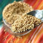 Spoonful of golden colored popped quinoa with bowl of toasted quinoa in background, sitting on top of red, orange, and blue cloth napkin.