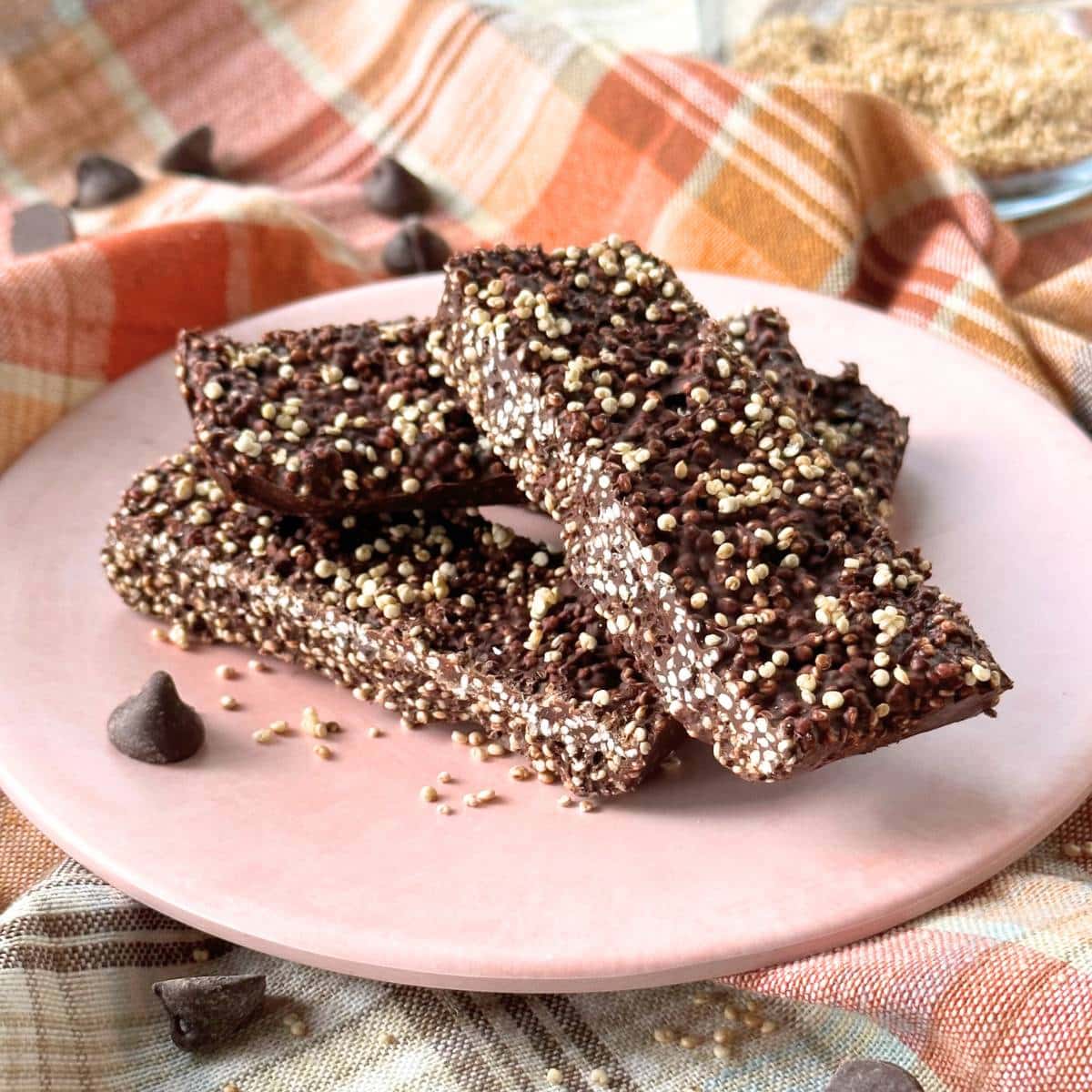 Picture of chocolate quinoa crunch bar using popped quinoa. Chocolate quinoa crunch bars are stacked on a pink plate on top of a plaid colorful cloth. Background shows a bowl of popped quinoa with popped quinoa and chocolate chips scattered around.