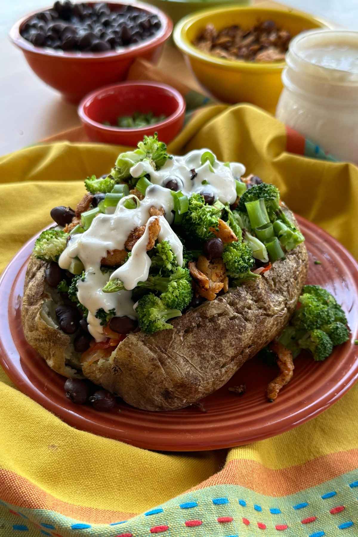 Stuffed baked potato with black beans, broccoli, soy curls, and green onions with aioli drizzled on top. On red plate on top of yellow, orange, and green cloth napkin with blue and red stitching. Ingredients in background in serving dishes.