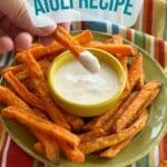 Sweet potato fry dipped in aioli with aioli in dipping sauce bowl and sweet potato fries on green plate in background on top of striped colorful cloth napkin. Text overlayed on top reads "Vegan AIOLI RECIPE" and "AlternativeDish.com"