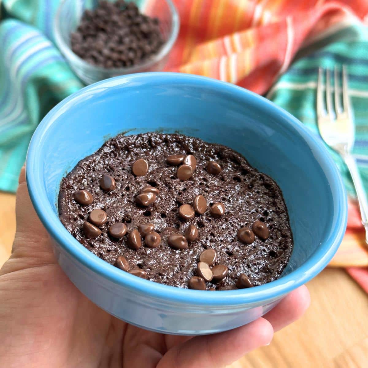 Vegan individual microwave brownie in a small 8 ounce blue bowl. Hand holding the bowl to show shallow depth. Colorful striped cloth in background with fork and bowl of chocolate chips.
