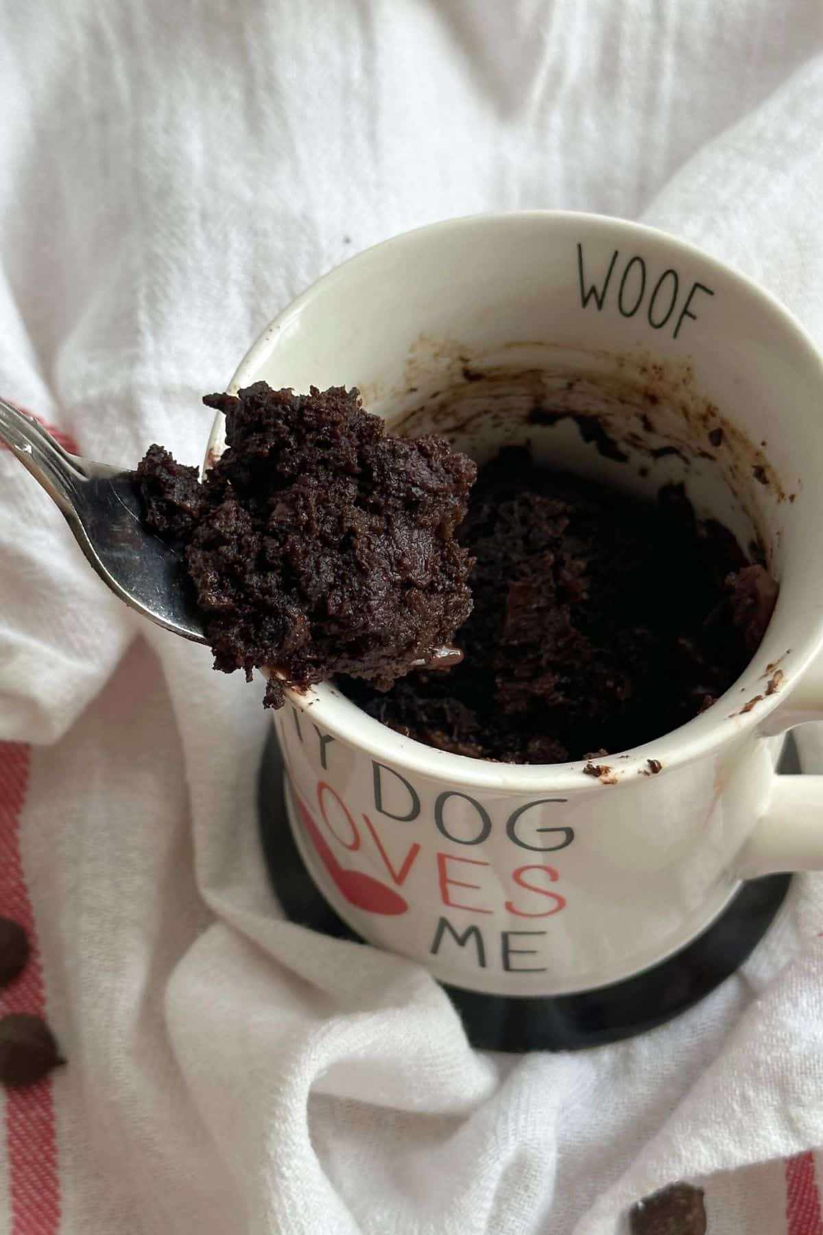 Bite of chocolate zucchini cake on fork lifted out of mug with more cake. White mug with text that reads "WOOF" on lip. On top of white cloth napkin with red stripes.