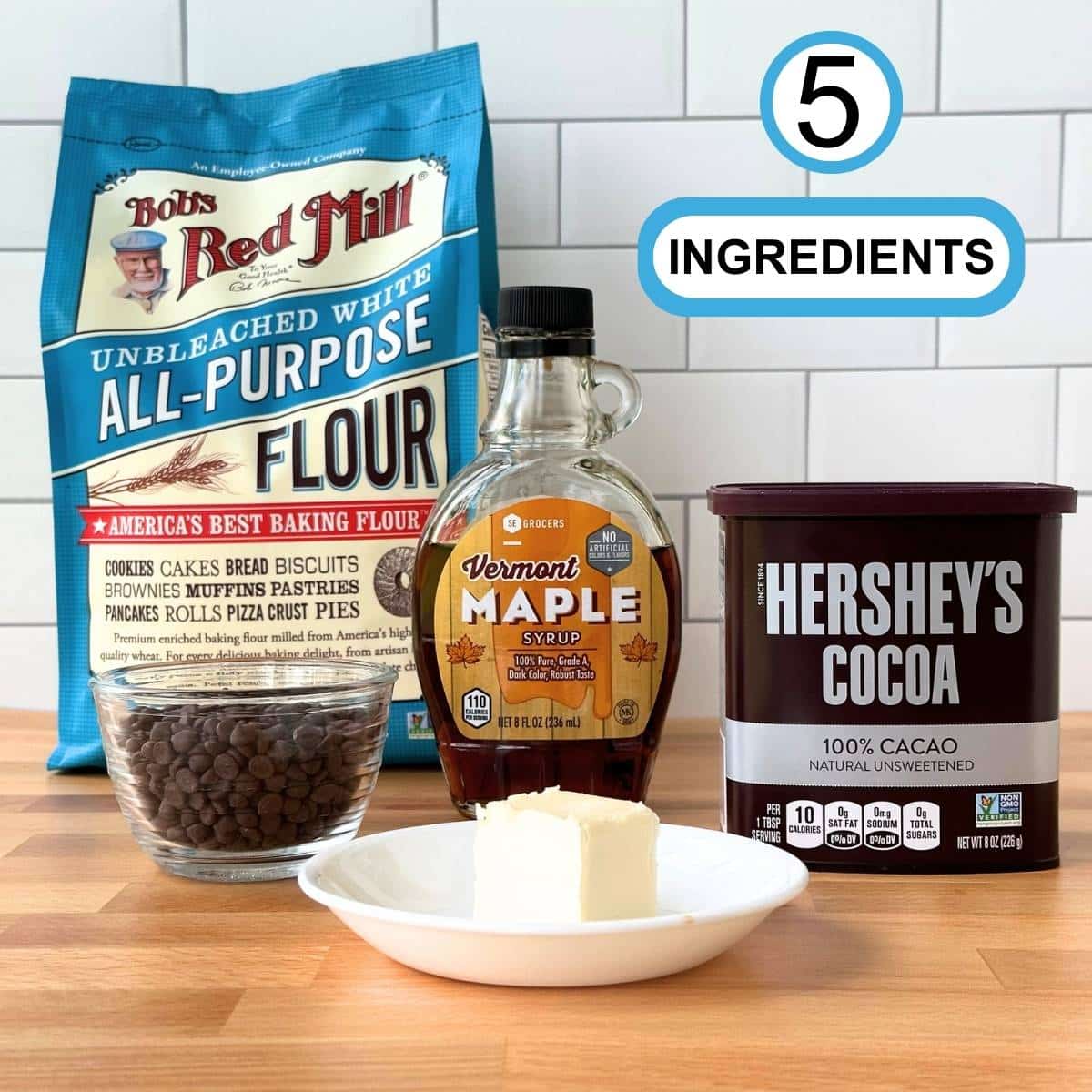 Picture of ingredients for vegan mug brownie recipe: Bag of all purpose flour, bowl of mini chocolate chips, bottle of maple syrup, cocoa powder, and stick of butter on white plate. Text reads "5 INGREDIENTS" in black on white bubble with blue border.