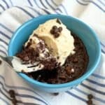 Vegan mug brownie in blue bowl with ice cream on top. Fork lifting out bite of mug brownie with ice cream. Background is white cloth napkin with thin blue stripes and chocolate chips sprinkled on top.