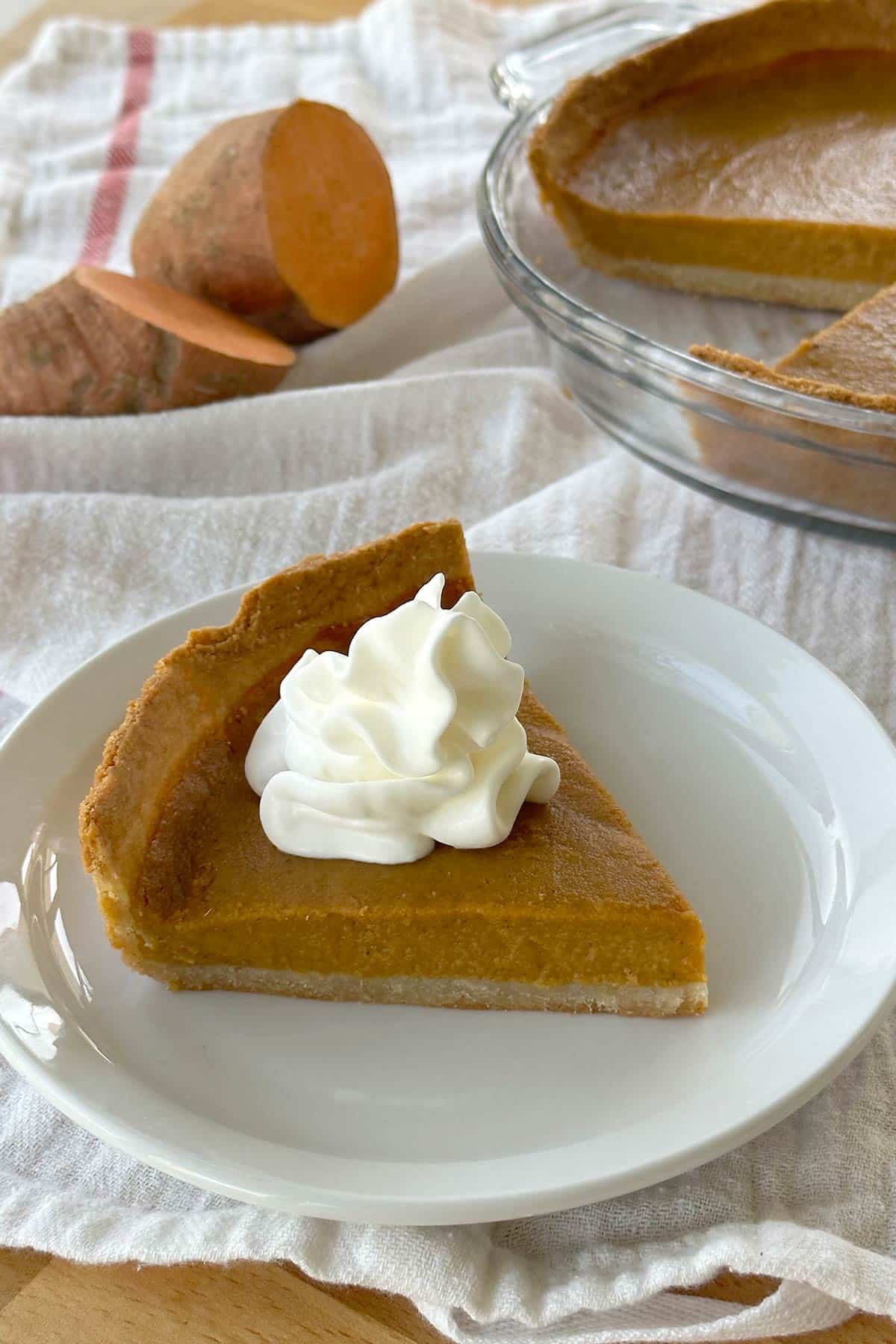 Slice of vegan sweet potato pie with dollop of whipped cream on top of a white plate. Background shows pie plate with the rest of the sweet potato pie in it and a cut open sweet potato.