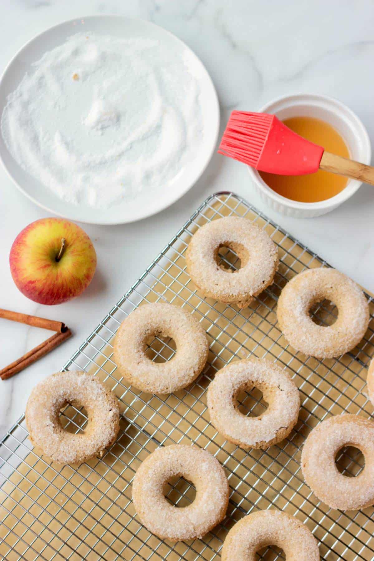 Baked apple cider donuts on a wire rack with sugar coating. Background shows the bowl of sugar and apple cider for sugar coating process along with an apple and 2 cinnamon sticks.