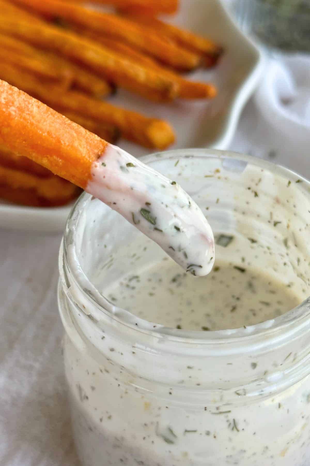 Sweet potato fry dipped in vegan ranch dressing. Glass jar filled with vegan ranch dressing, plate with sweet potato fries, and dairy-free ranch seasoning are in the background on top of a white cloth.