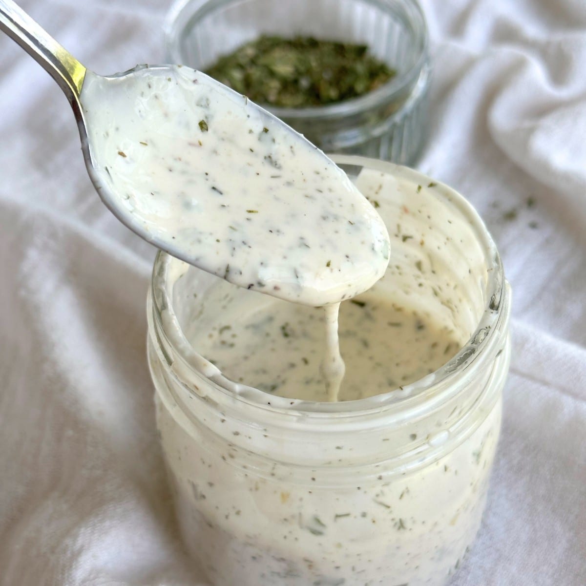 Spoonful of vegan ranch dressing dripping into a glass mason jar full of vegan ranch dressing. Bowl of dairy-free ranch seasoning is in the background on top of a white cloth.