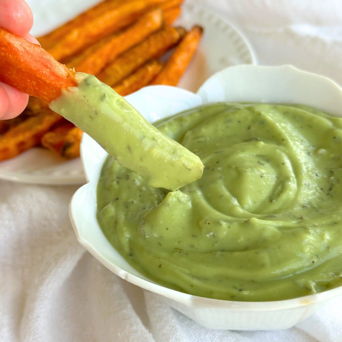 Sweet potato french fry dipped in vegan avocado ranch dip. background shows a plate of sweet potato fries.