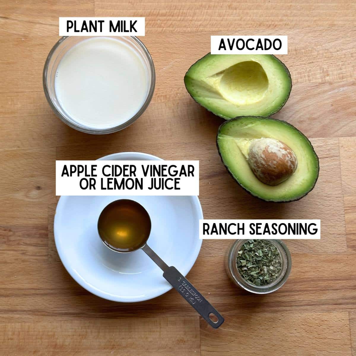 Ingredients to make vegan avocado ranch dressing: plant milk (in a glass bowl), avocado (sliced open in two halves), apple cider vinegar or lemon juice (in a tablespoon on top of a white plate), and ranch seasoning (in a glass bowl).