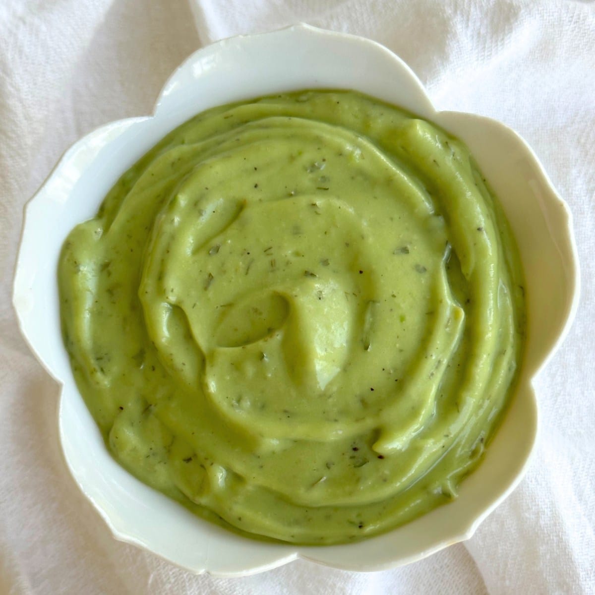 Bowl of vegan avocado ranch dressing with flecks of dairy-free ranch seasoning in creamy green sauce. Background is a white cloth.