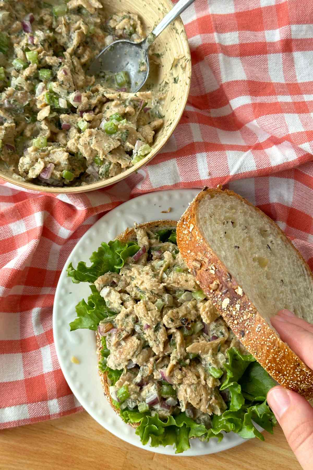 Vegan chicken salad on a sandwich with multigrain bread and large piece of lettuce underneath. The top slice of bread is being lifted up. In the background is the bow of vegan chicken salad on top of a red gingham napkin.