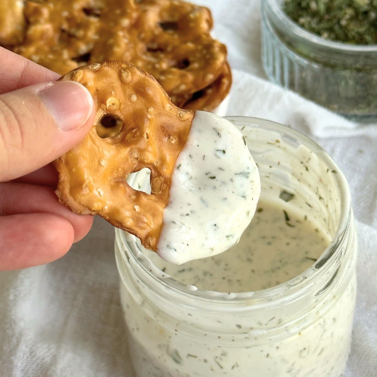 Flat pretzel chip dipped in vegan ranch dressing. Glass jar filled with vegan ranch dressing, plate with pretzels, and dairy-free ranch seasoning are in the background on top of a white cloth.