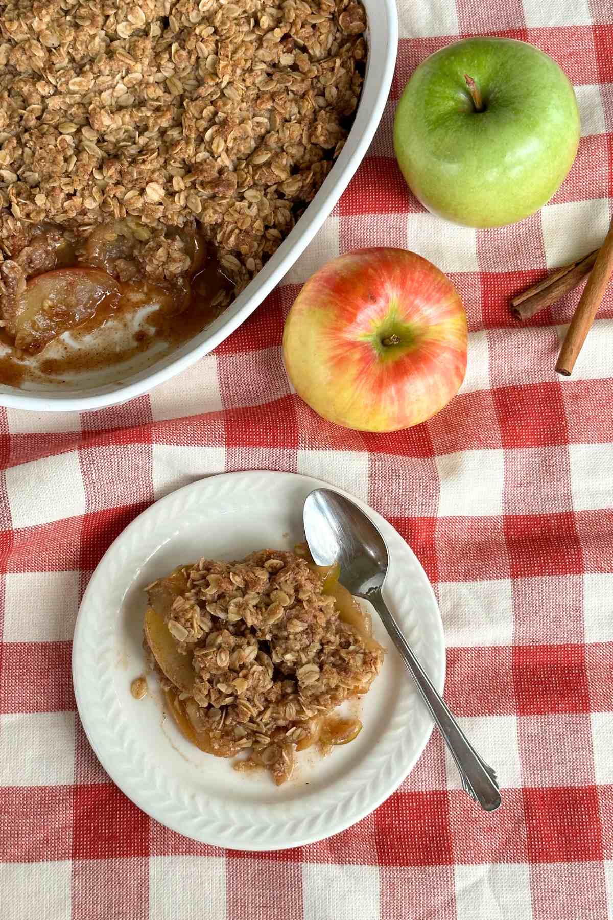 Plate with vegan apple crisp and a spoon next to the dish of apple crisp, a red apple, green apple, and two sticks of cinnamon, all on top of a red and white gingham napkin.