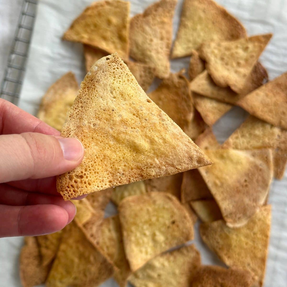 Chickpea tortilla chip being held with more chickpea chips on a parchment sheet in the background.