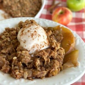 Dairy free apple crisp in a bowl with a scoop of vegan vanilla ice cream on top with cinnamon sprinkled over. The background shows the dish of vegan apple crisp, a red apple, green apple, and two sticks of cinnamon.