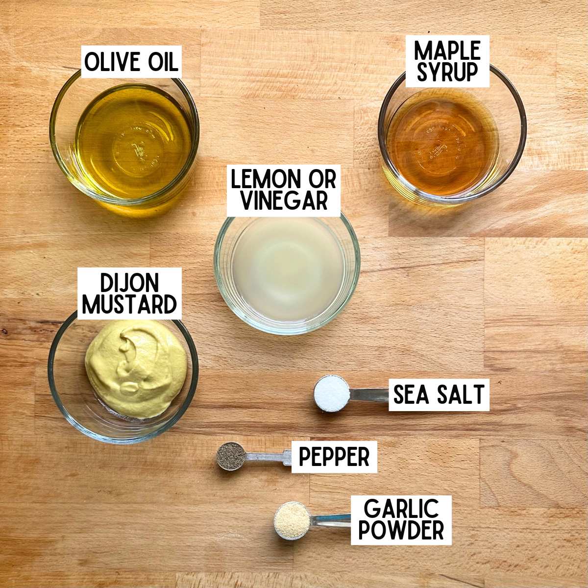 Ingredients to make maple Dijon vinaigrette with corresponding labels: olive oil, maple syrup, lemon or vinegar, Dijon mustard, sea salt, pepper, garlic powder.