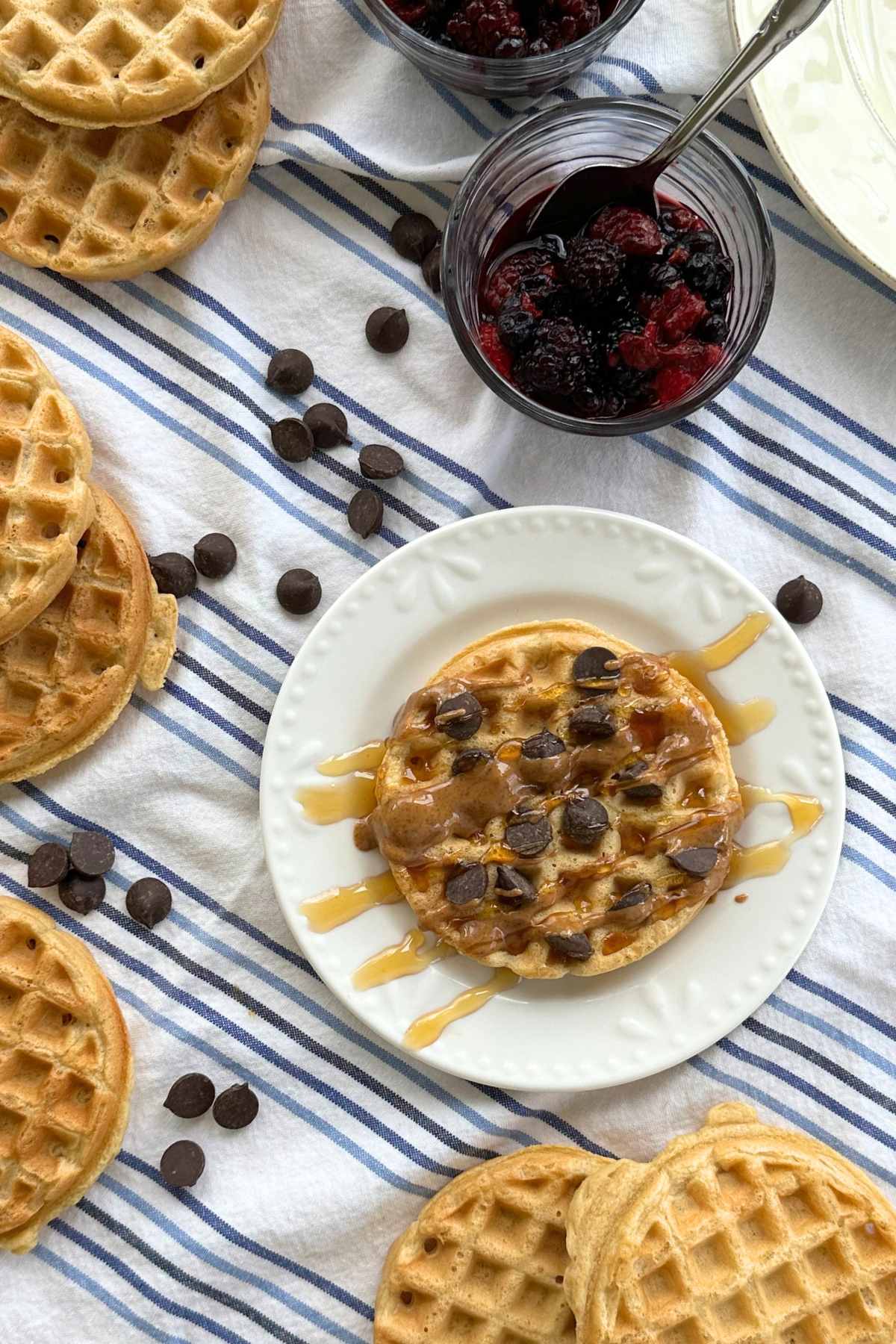 Protein waffle with almond butter and maple syrup drizzled over it and chocolate chips sprinkled on top. Around the plate with the waffle are more waffles, chocolate chips, and a bowl of mixed berries on top of a blue and white striped napkin.