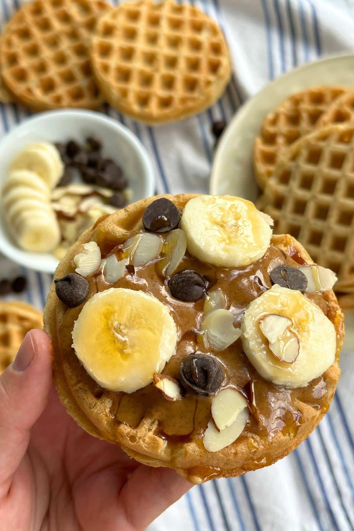 Vegan protein waffle with almond butter, banana slices, chocolate chips, slivered almonds, and maple syrup. background shows more waffles and a bowl with the toppings in it on top of a white and blue striped cloth napkin.