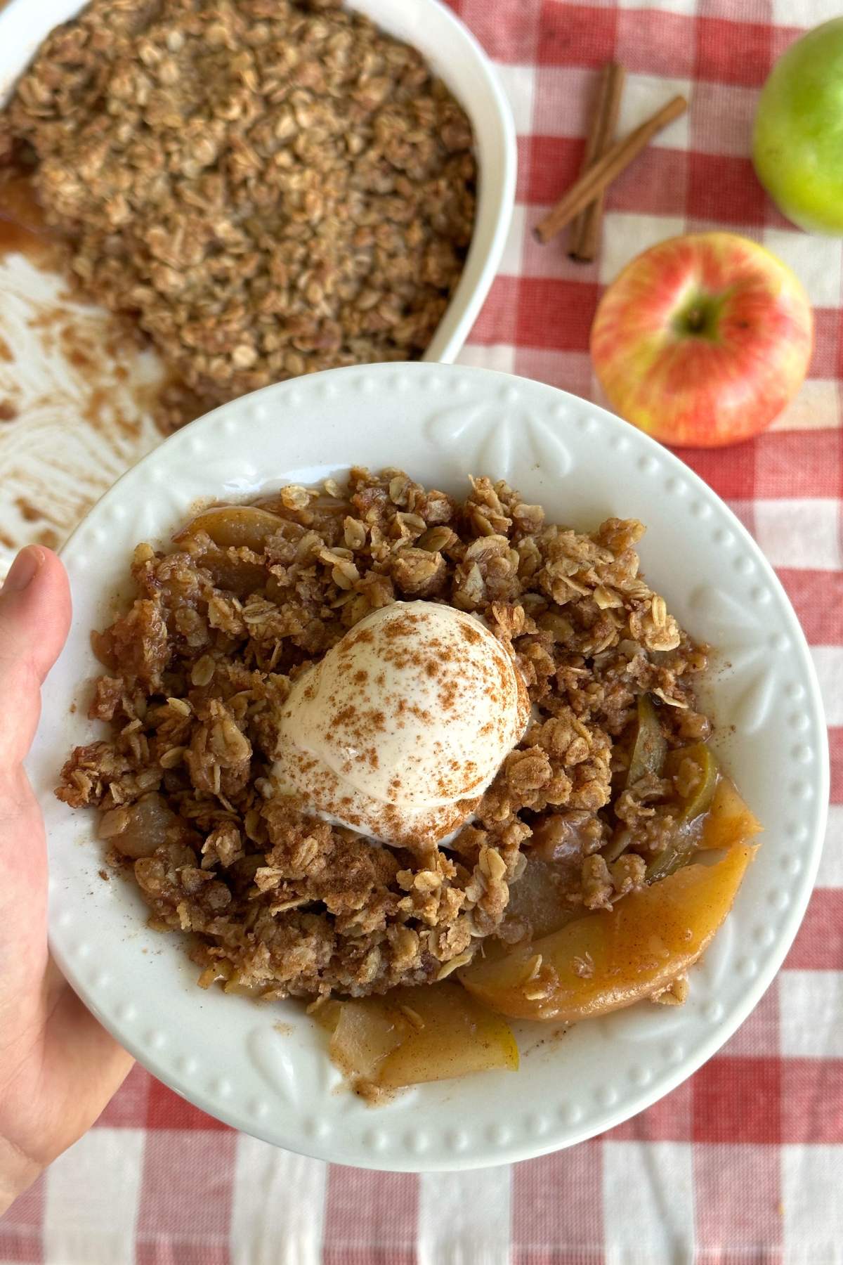 Vegan apple crisp in a bowl with a scoop of vegan vanilla ice cream on top with cinnamon sprinkled over. The background shows the dish of vegan apple crisp, a red apple, green apple, and two sticks of cinnamon.