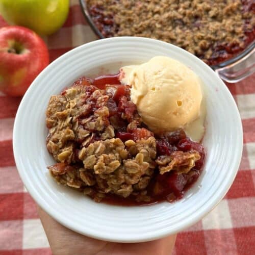 Vegan cranberry apple crisp in a bowl with a scoop of ice cream. The background shows a red and green apple and the crisp in a baking dish on top of a red and white gingham napkin.