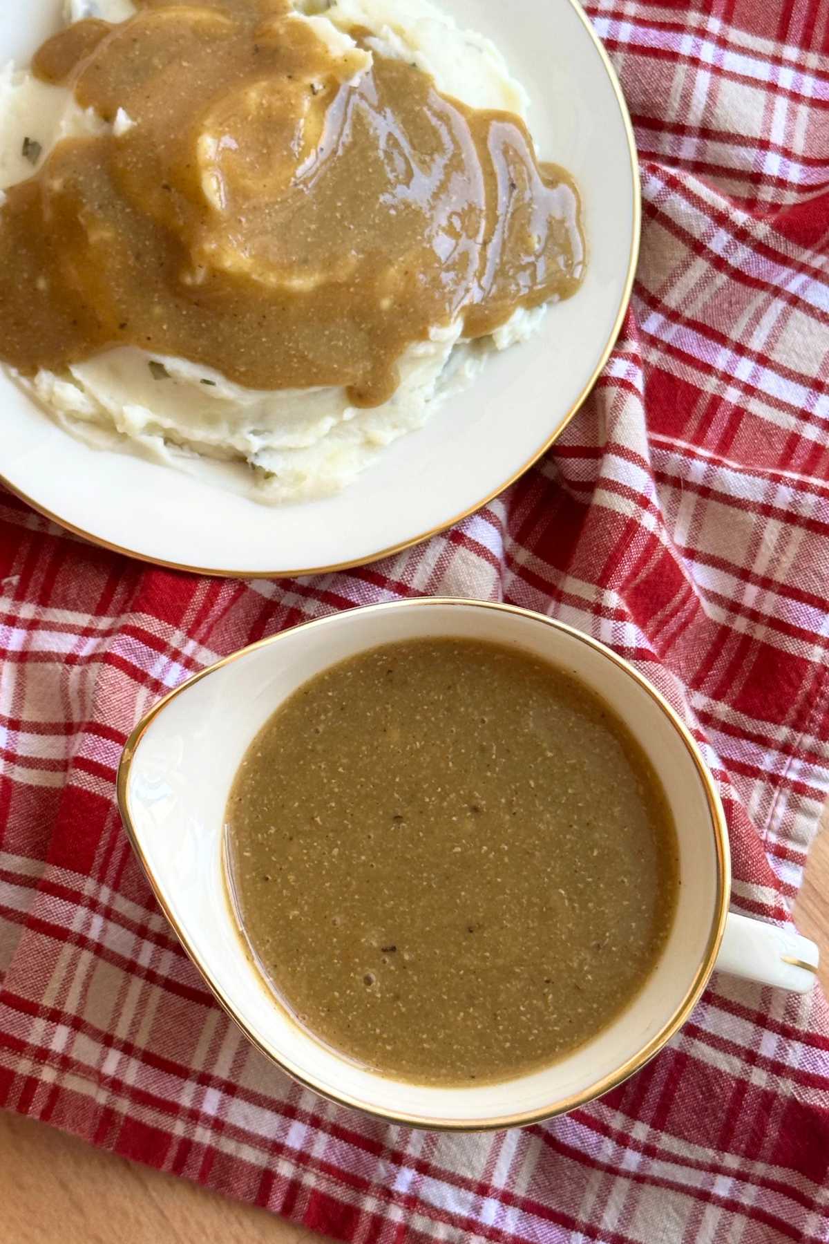 Overhead photo of plate of mashed potatoes with gravy poured on top and the sauce dish with gravy in it, both are resting on top of a red and white plaid napkin. 