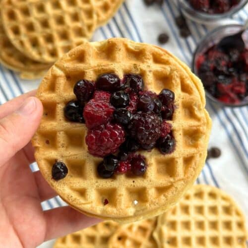 Vegan protein powder waffle with mixed berries piled on top. The background shows more waffles and the bowl of berries on a white and blue striped cloth napkin.