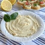 Vegan tofu ricotta in a white bowl with two basil leaves on top. A lemon is sliced in half in the background along with a plate of crostinis.
