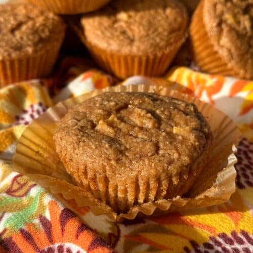 Apple muffin with wrapper pulled down. Background shows more apple muffins piled on top of colorful fall flower napkin.