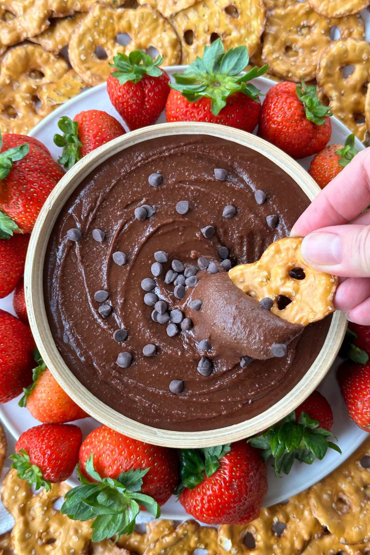 Pretzel being dipped into brownie batter hummus with chocolate chips. Strawberries and pretzel chips surround the bowl of hummus.