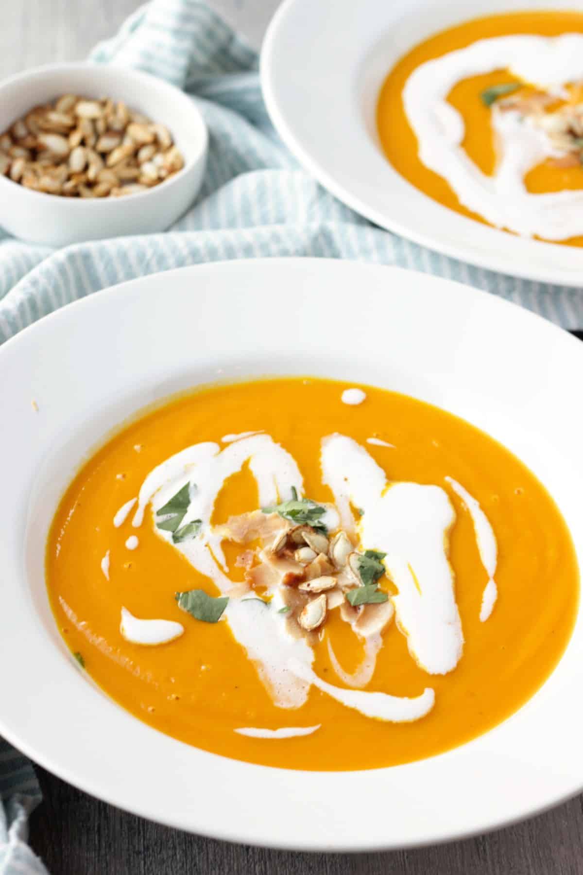 Two bowls of butternut squash soup with coconut cream, cilantro, and roasted squash seeds taken in perspective. A small bowl of squash seeds and 2 spoons are on top of a white and blue striped cloth and wood background.