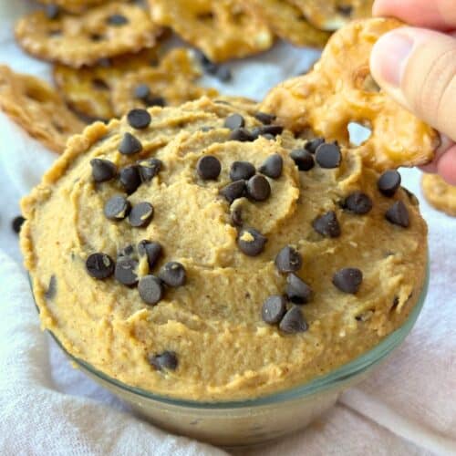 Chickpea cookie dough dip in a bowl with more chocolate chips sprinkled over top. A pretzel chip is being dipped into it.