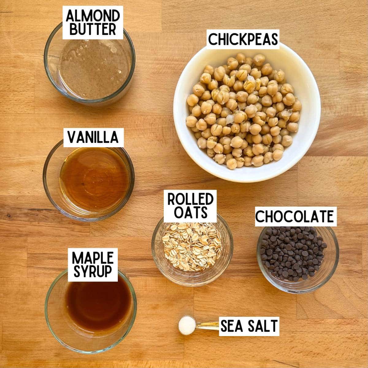 Ingredients to make chickpea cookie dough with corresponding labels: almond butter, chickpeas, vanilla, rolled oats, chocolate, maple syrup, and sea salt.