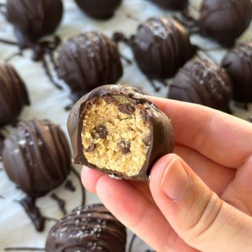 Chocolate covered chickpea cookie dough truffle with a bit taken out of it. The background shows more truffles.
