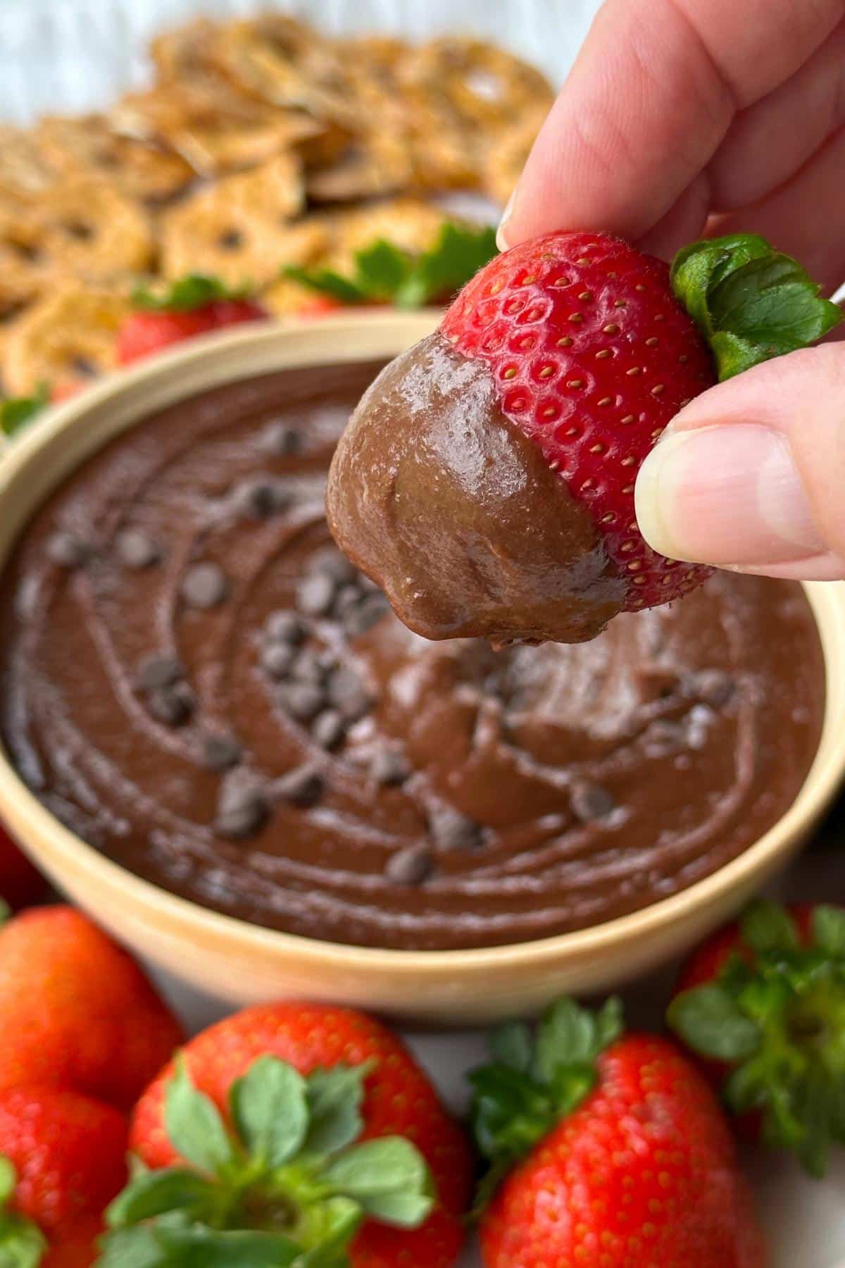Strawberry dipped in brownie batter dessert hummus. The bowl of hummus, strawberries, and pretzel chips are in the background.