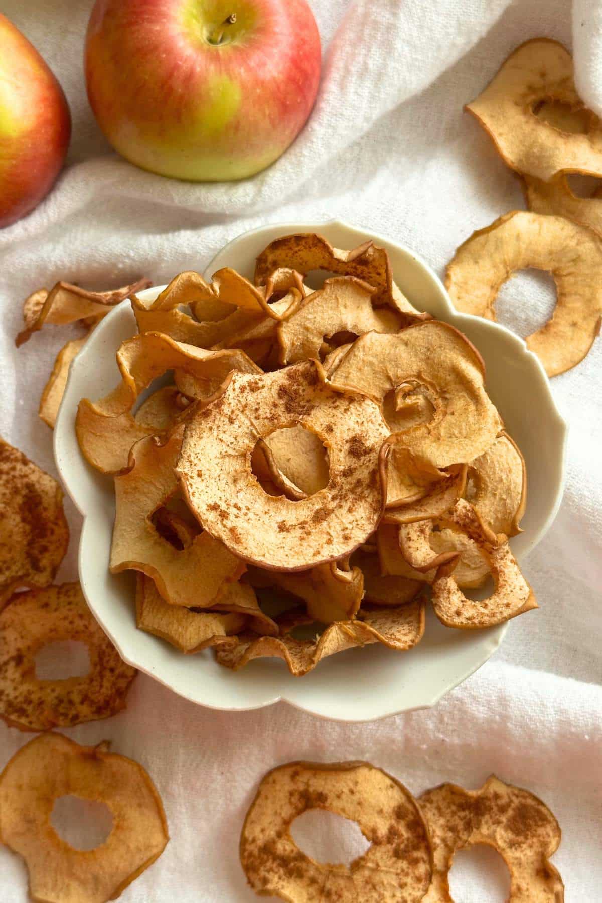 Baked apple chips in a bowl with more apple chips scattered around in the background with 2 apples.