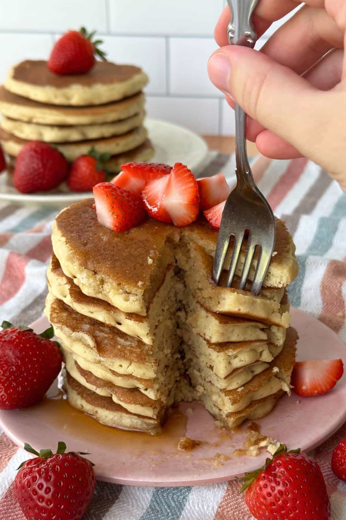 Stack of pancakes with a piece cut out and the fork holding it. Strawberries are on top and around the pancakes.