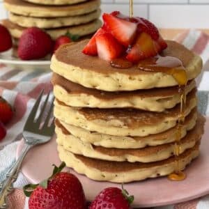 Vegan Bisquick pancakes stacked on a pink plate with chopped strawberries piled on top and maple syrup being poured. More pancakes and strawberries are in the background.